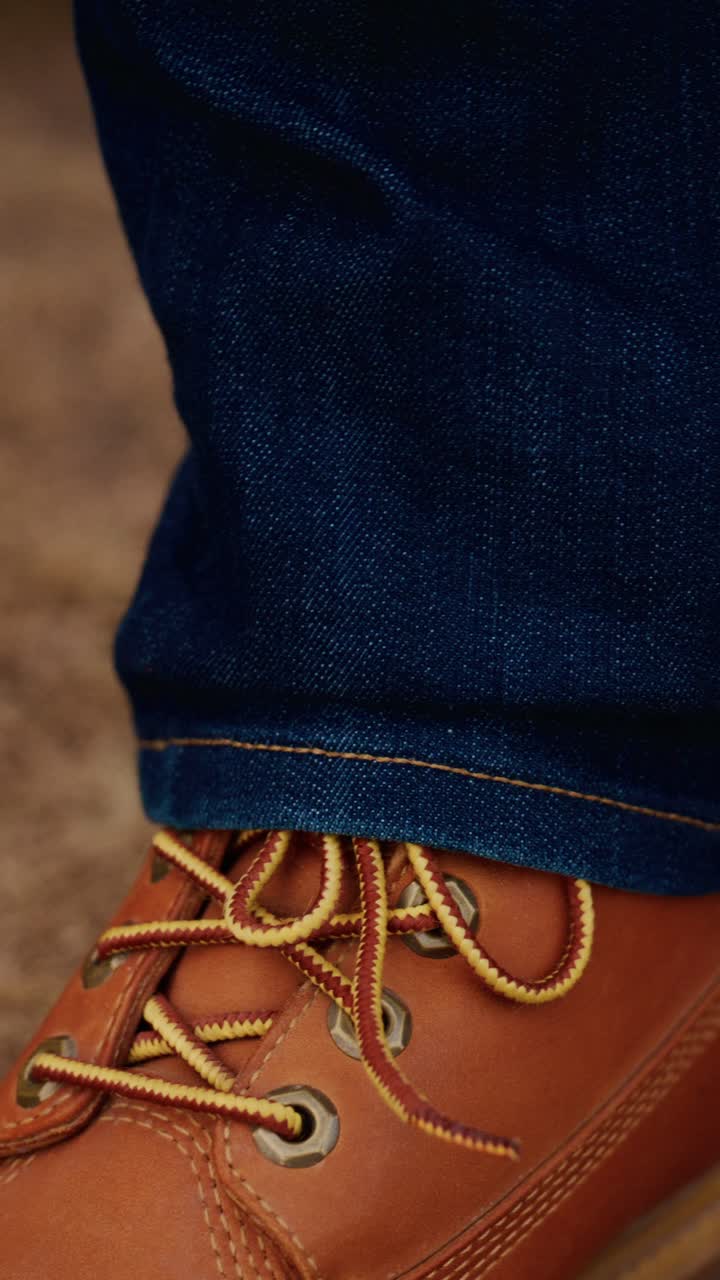 Close-Up Detail Tangan Mengikat Tali pada Sepatu Bot Kulit Cokelat, Menampilkan Tekstur dan Keahlian dalam Suasana Kasual dengan Penekanan pada Alas Kaki dan Fashion