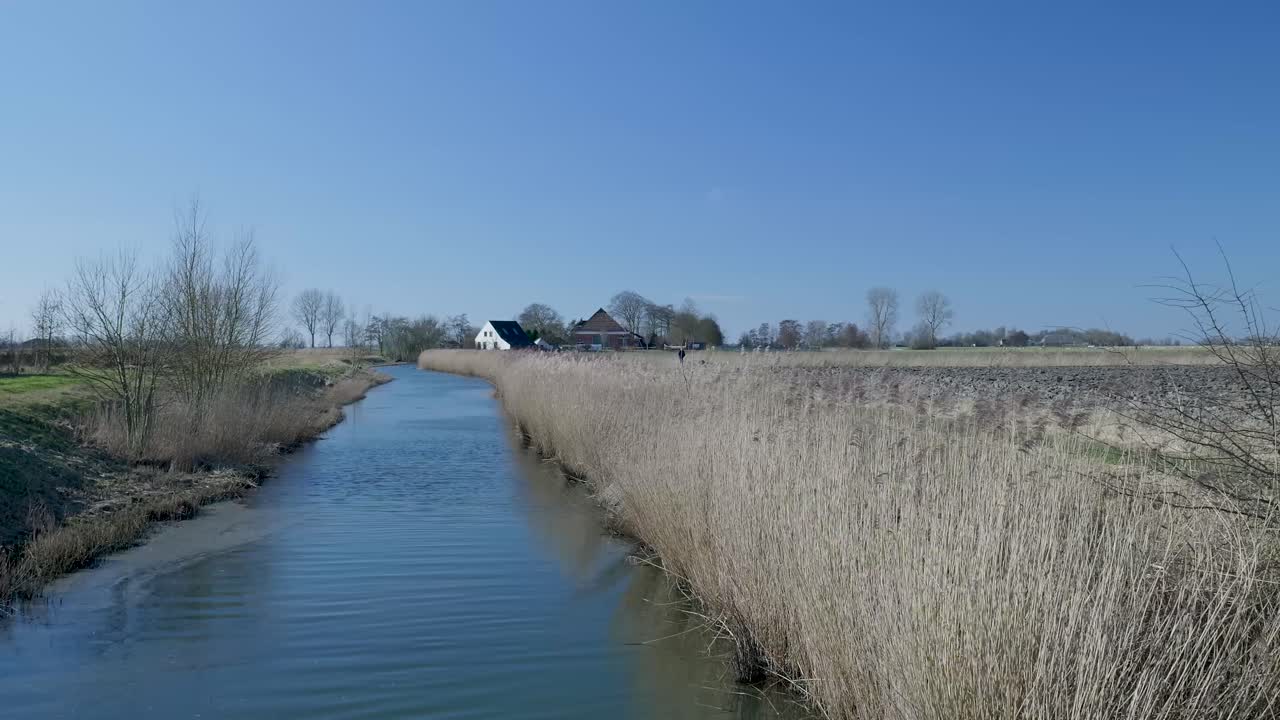 Fredfyldt landskab med kanal, siv og fjerne landhuse under blå himmel