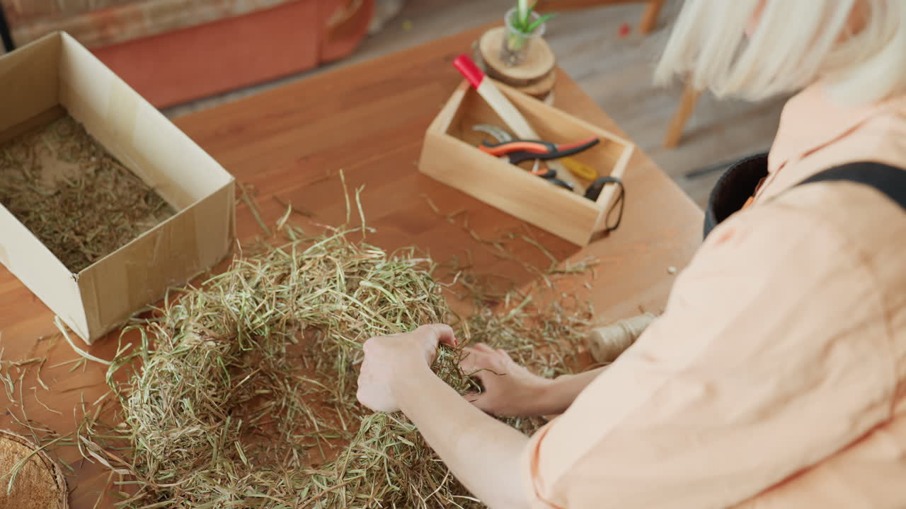 Person shaping hay wreath on wooden table with tools and cardboard box nearby, wearing apron and peach shirt, crafting natural handmade rustic decoration with eco-friendly materials