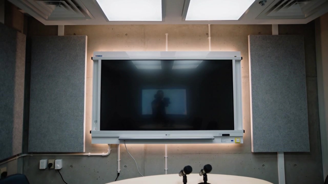 Mostrando una pantalla grande que refleja a una persona acercándose, levantando el teléfono en una sala de reuniones, probando micrófonos.