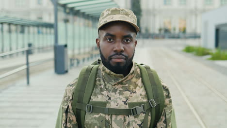 Portrait-of-serious-African-American-handsome-young-male-soldier-in-cap-with-backpack-standing-at-street.-Man-militarian-looking-at-camera-at-train-station.-Bus-stop.-Military-uniform.-Zooming-in.