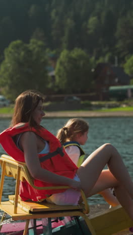 mother and daughter pedale catamaran moving near shore. river with lush green vegetation and tourist lodges side view. woman and child rest enjoying movement on water slow motion