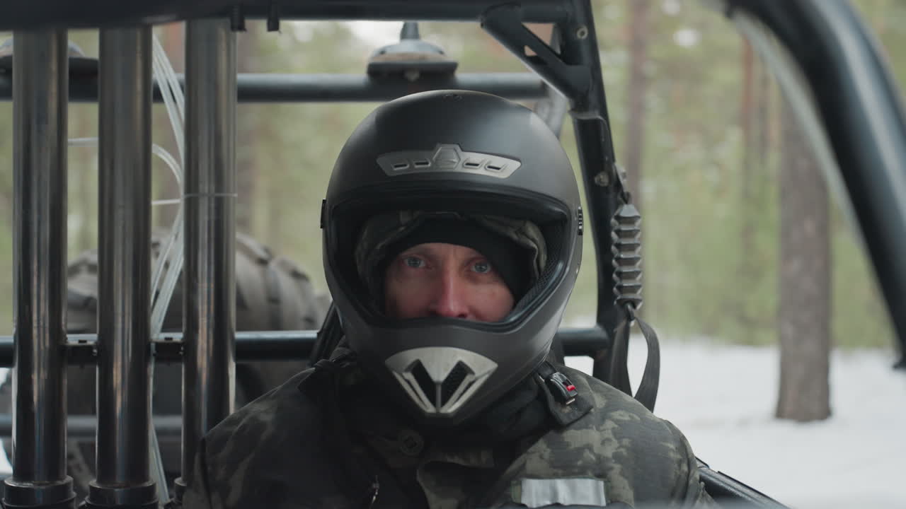 Teknisi pria berhelm di kabin ATV memeriksa kontrol dan instrumen, tangan bersarung di dasbor, radio terjepit di jaket, roll cage tertutup salju tipis, inspeksi tenang sebelum uji performa