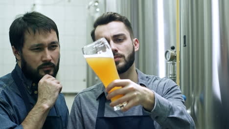 two male coworkers smiling, looking at freshly brewed unfiltered beer in glass and discussing it at brewery