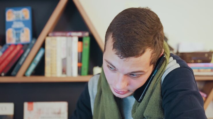 Studente caucasico pensieroso alla scrivania con libreria, primo piano, con sciarpa verde, illuminazione soffusa e maglione invernale accogliente, che studia e prende appunti, espressione introspettiva, atmosfera tranquilla da biblioteca domestica, concentrato.