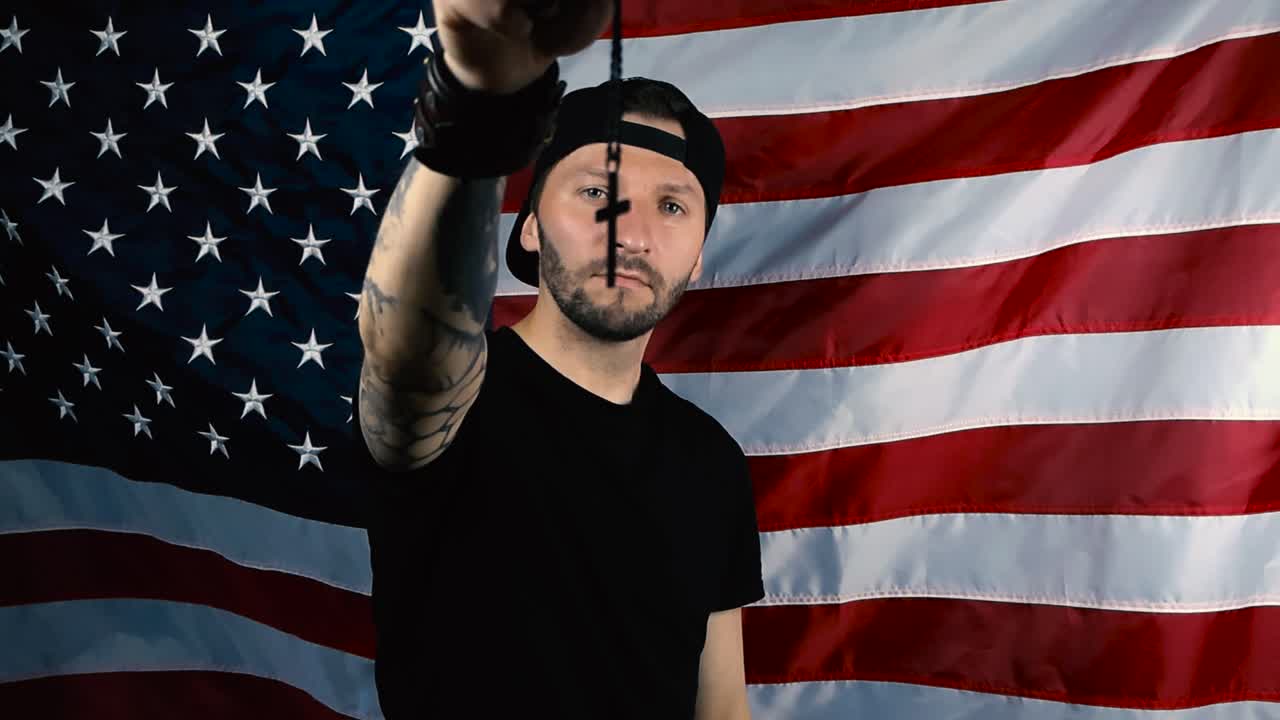 Handsome tattooed man lifts up a black colored holy cross on a chain with his hand in front of an American United States flag and nods with confidence. Holy person and flag moving in slow motion