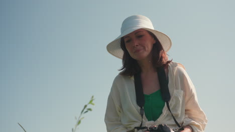 tourist in wide-brimmed white hat and cream shirt holding camera while crouched in green field under clear sky, preparing to photograph flower with focused expression on warm sunny day