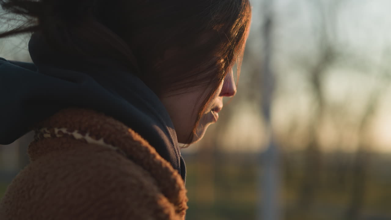 해가 지는 동안 공원을 걸어다니는 눈물을 흘리는 소녀, 갈색 코트와 후디를 입고, 클로즈업 은 그녀의 감정적 상태를 강조하고, 부드럽고 황금색 빛이 그녀의 얼굴을 가라앉힌다.