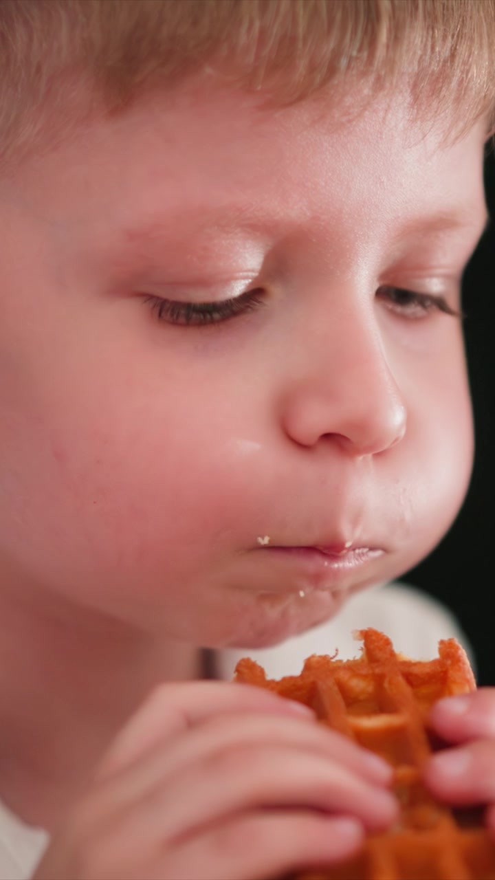 Close up of young boy eating waffle. child holds and takes bites from food with focus on his hands holding it. Background is dark emphasizing subject and action