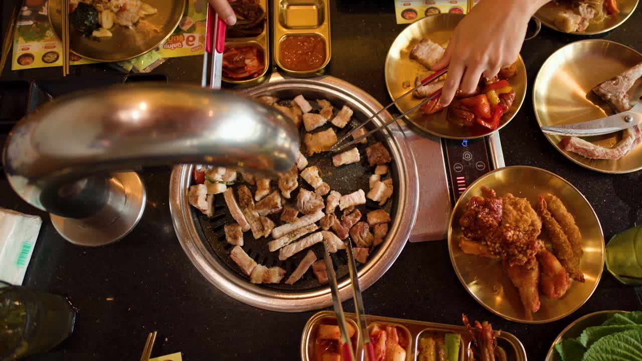 Overhead view of hands grilling pork belly with tongs, surrounded by Asian dishes and sauces