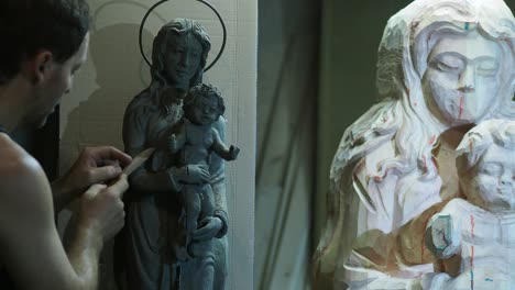 adult fit sculptor carefully adjusts details on a small clay model of a madonna and child statue, using it as a guide while working on the larger wooden version beside it in studio in lombardy, italy