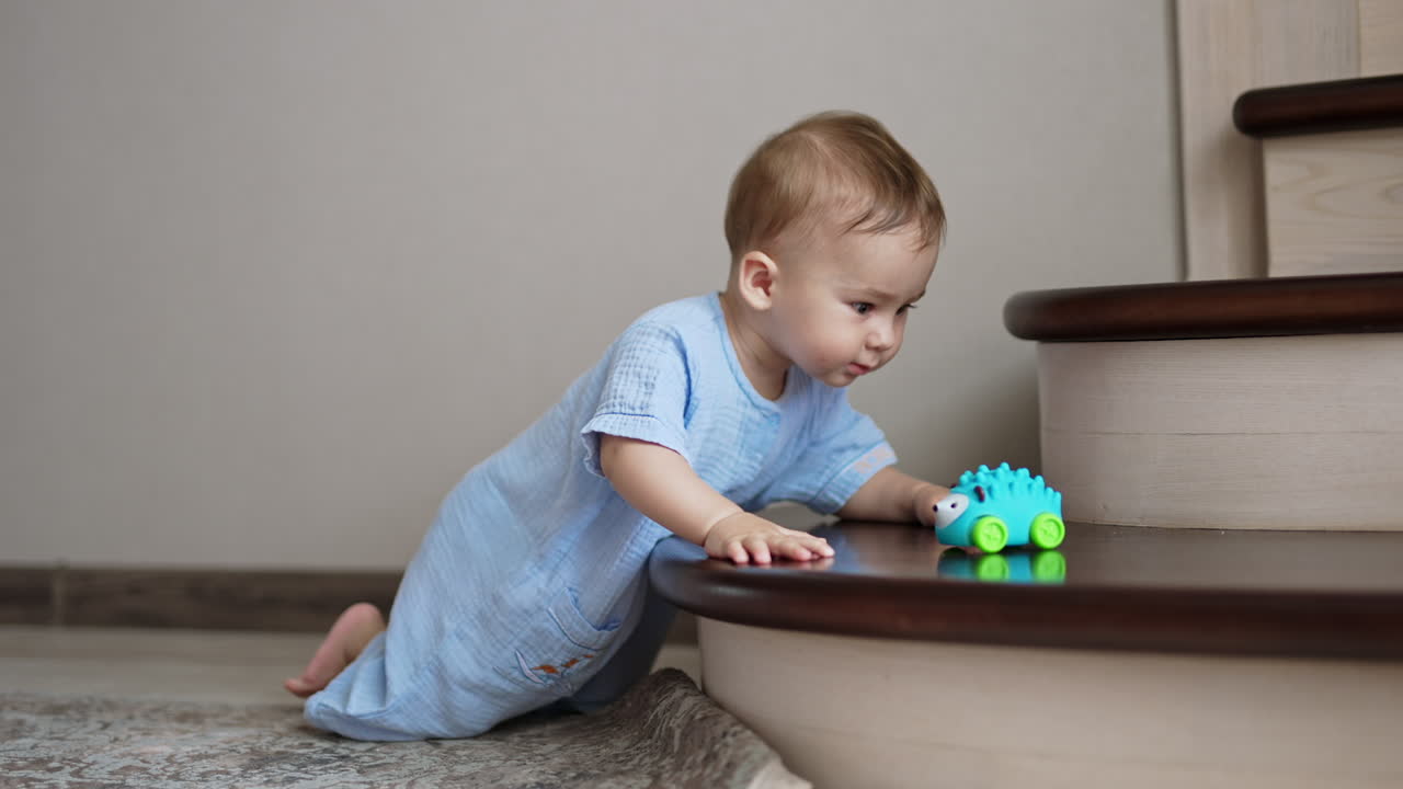 Happy smiley baby boy standing on his knees near the stairs inside the house. Adorable kid looks at toy and sways cheerfully.