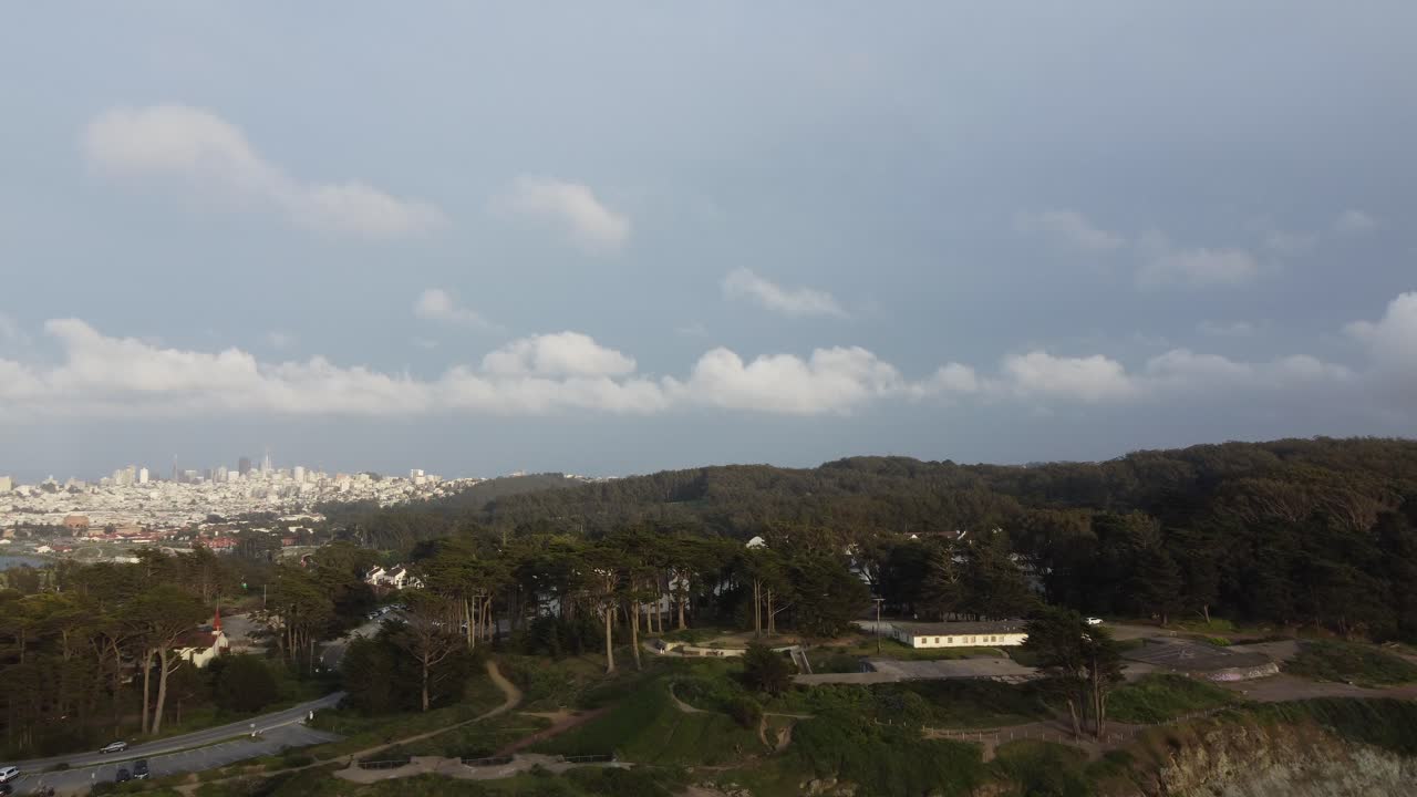 The San Francisco Presidio: Slow Aerial Ascend Over Coastal Cliffs And Tree Covered Hillside. The Historic Park Offers Stunning Views Of The Downtown City Skyline To The Far Left Of The Frame.