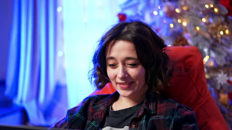 Cozy night with my laptop. A young woman relaxes in a red chair, using her laptop in a warm and festive atmosphere with lights and decorations