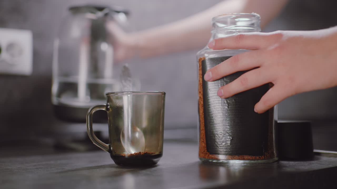 Close up of person scooping coffee from glass jar into mug while standing by kitchen counter, preparing hot drink, showing hands holding spoon and container beside electric kettle in natural daylight