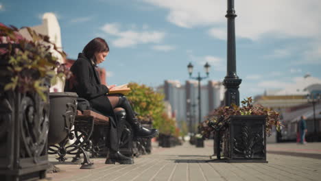 side view of stylish woman seated in public space flipping through book, flowers swaying gently with wind, pedestrians walking, stores with inscription across road in background and street lamp