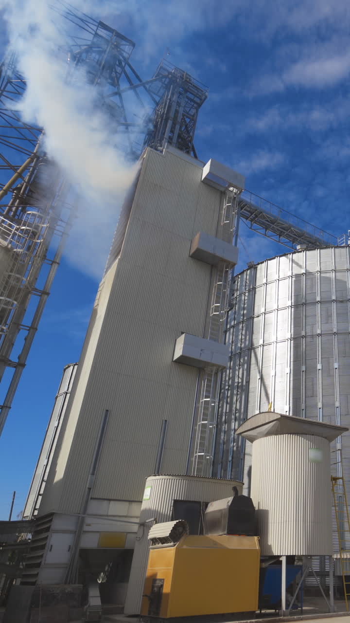 Agricultural Silos. Metal grain facility with silos. Storage and drying of grains, wheat, corn, soy, sunflower against the blue sky with white clouds Vertical video