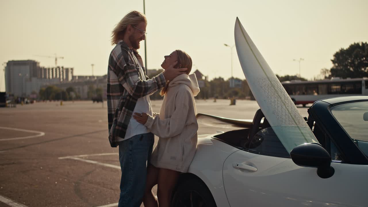 Close-up shot of a blond guy with glasses with a beard in a plaid shirt hugs his blonde girlfriend and strokes her hair, a girl in a white sweatshirt looks at her boyfriend and leans on a white convertible in which there is a white surfboard on a large square in the city