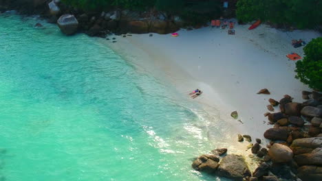 una toma aérea de una playa de arena blanca con piedras grandes, agua azul clara y gente irreconocible en la arena