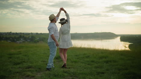 a gentle man twirls his wife around on a grassy hill beside a tranquil lake during a beautiful sunset. the man, dressed in a white shirt, hat, and jeans, creates a loving moment with his wife