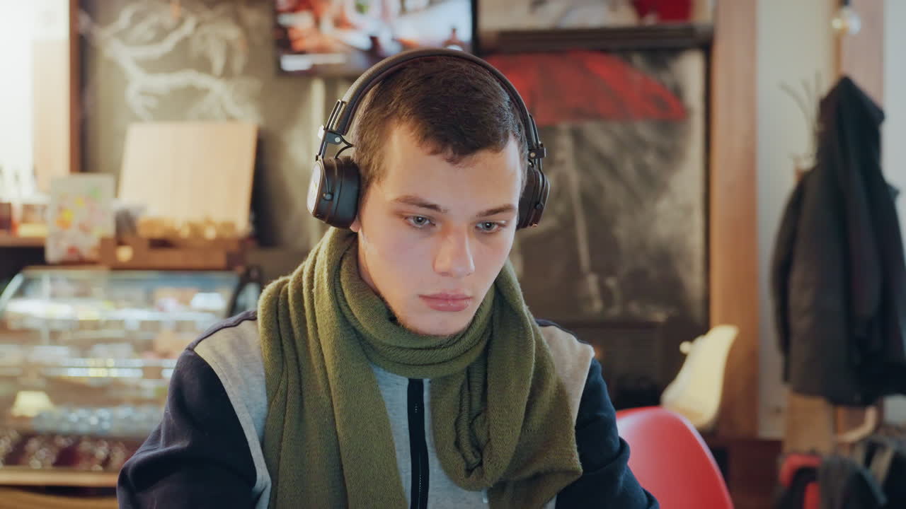 Kavkazský muž se sluchátky u notebooku, šála a teplé pozadí kavárny, klidný soustředěný pohled, pracovní postup řízený hudbou, gesta při úpravách a kódování, měkká světla, útulná atmosféra a stálá koncentrace.