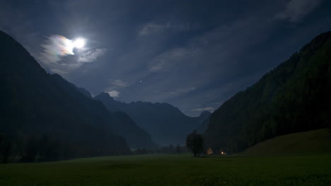 Timelapse-of-Alpine-valley-by-night-under-full-moon,-illuminated-farmhouse,-cottage,-full-moon,-clouds-and-stars-in-sky,-fog-in-the-valley