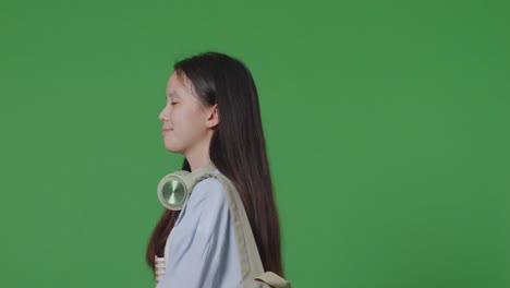 close up of side view asian teen girl student with a backpack holding some books and smiling while walking in the green screen background studio