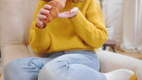 Una-Mujer-Casual-Come-Un-Delicioso-Croissant-En-El-Sofá,-Vista-Frontal