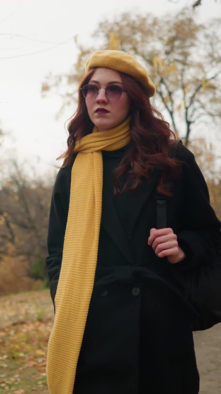 mujer con silenciador amarillo y boina camina por el bosque, ajustando gafas mientras sostiene la correa de la bolsa, rodeada de follaje de otoño