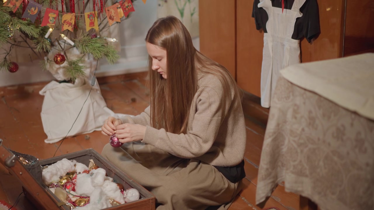 Scena festiva: una donna decora l'albero con un ornamento di vetro e un nastro, seduta accanto a un baule aperto pieno di palline e festoni, con sorrisi gentili e una calda tradizione familiare mentre la decoratrice sistema le luci.