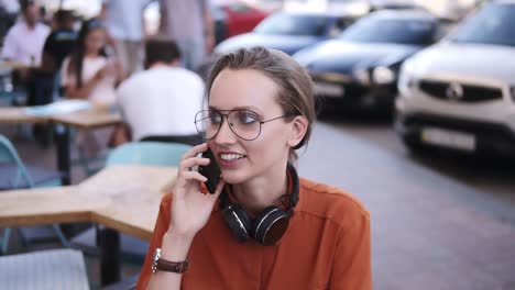 Close-up-of-fair-hair-girl-with-headphones-on-her-neck-talking-on-mobile-phone-while-sitting-in-street-cafe.-Smiling,-wearing