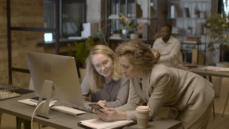 two female employees looking something at mobile phone and talking together in the office