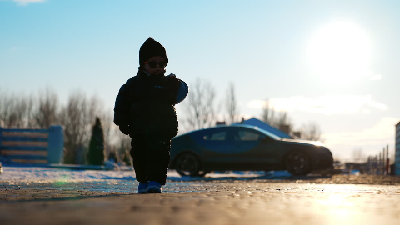 Lovely baby boy wearing black jacket, cap and sunglasses walks approaching camera. Cute kid carries keys from a car. Low angle view.