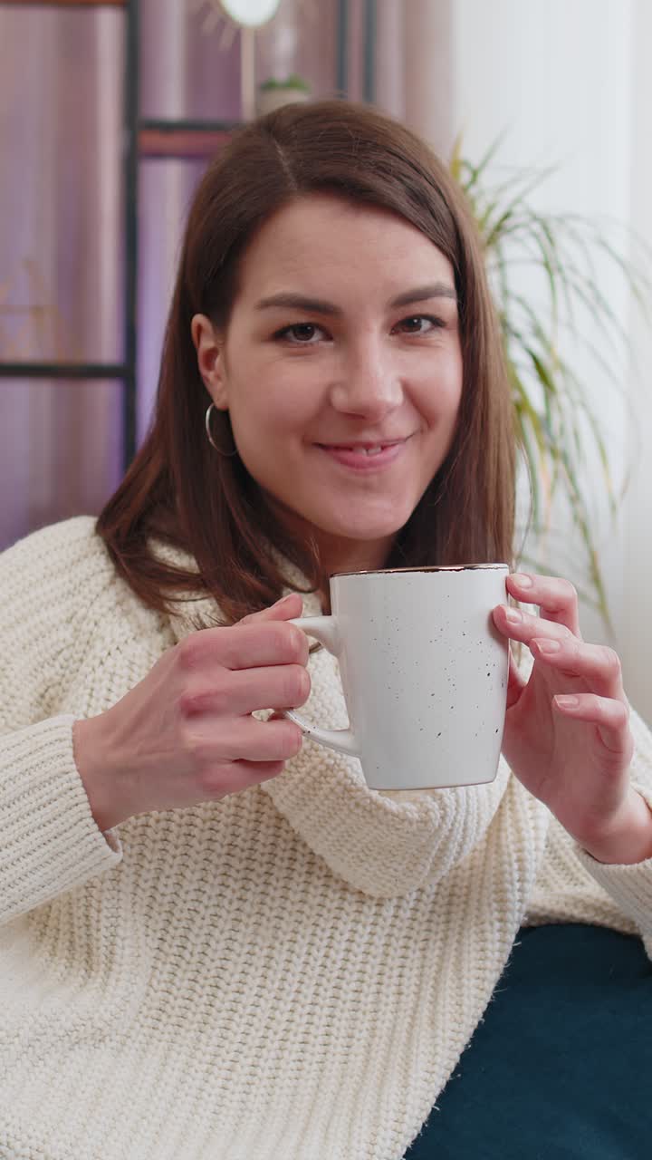 Affascinante giovane donna sorridente che beve una tazza di caffè o tisana sul divano di casa al mattino