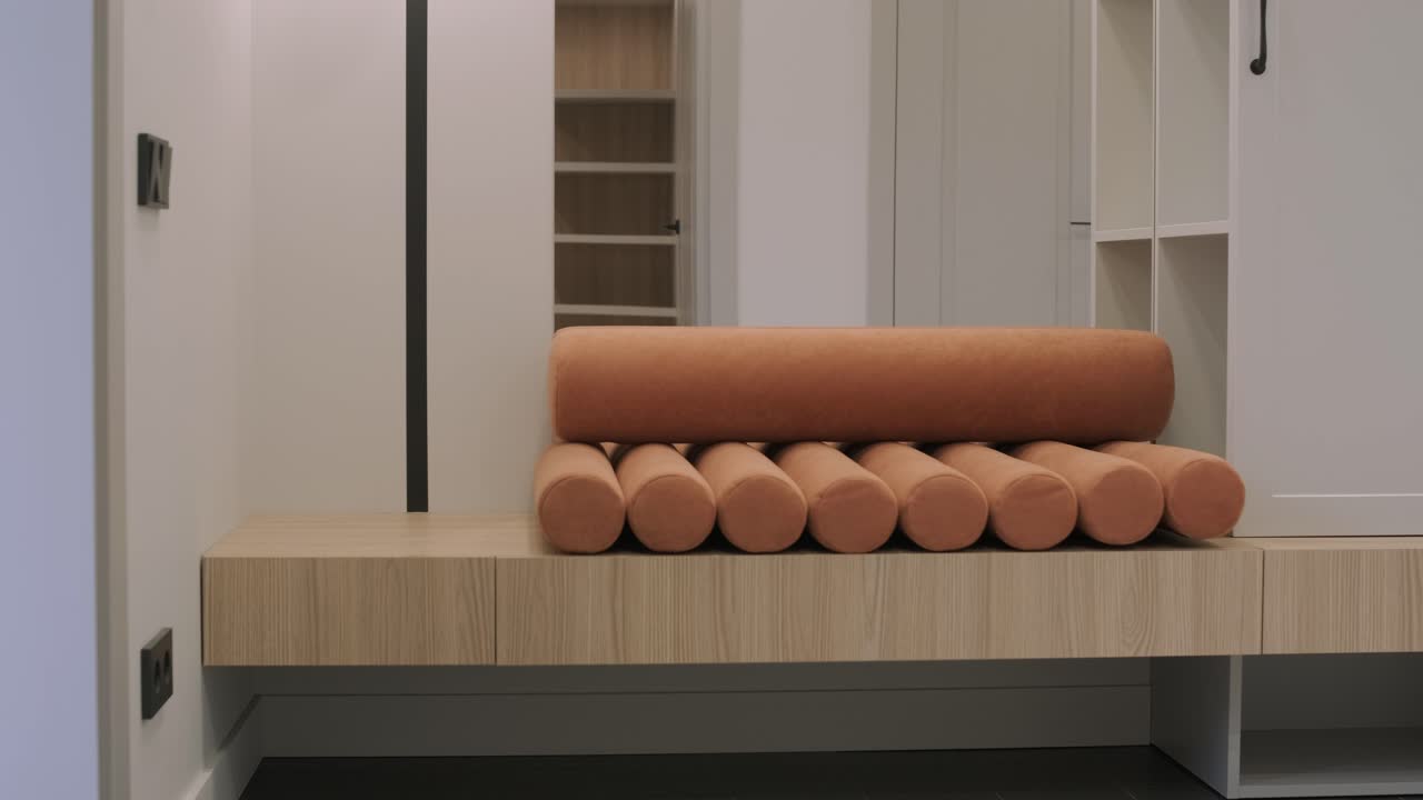 современный входный зал имеет минималистскую скамейку с уникальной цилиндрической подушкой, расположенную на фоне гладкого деревянного шкафа для обуви и нейтральной цветовой палитры, подчеркивая чистые линии и функциональный дизайн современного домашнего декора