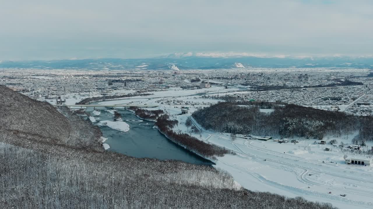 Aerial View Of Asahikawa City In Hokkaido, Japan With Ishikari River Flowing Through Snowy Mountain Landscape In Winter. wide shot