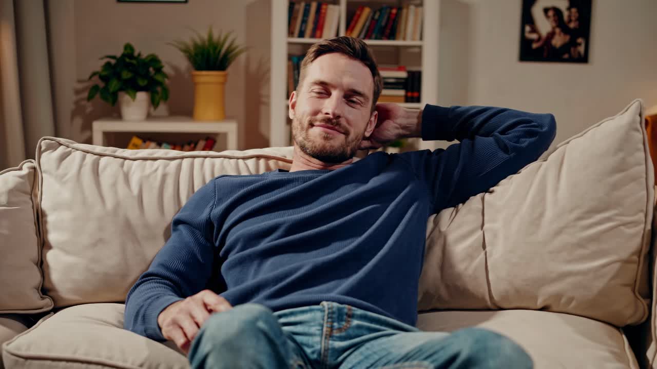 A relaxed man in a blue sweater lounges on a beige sofa, viewed from a low angle, creating a cozy