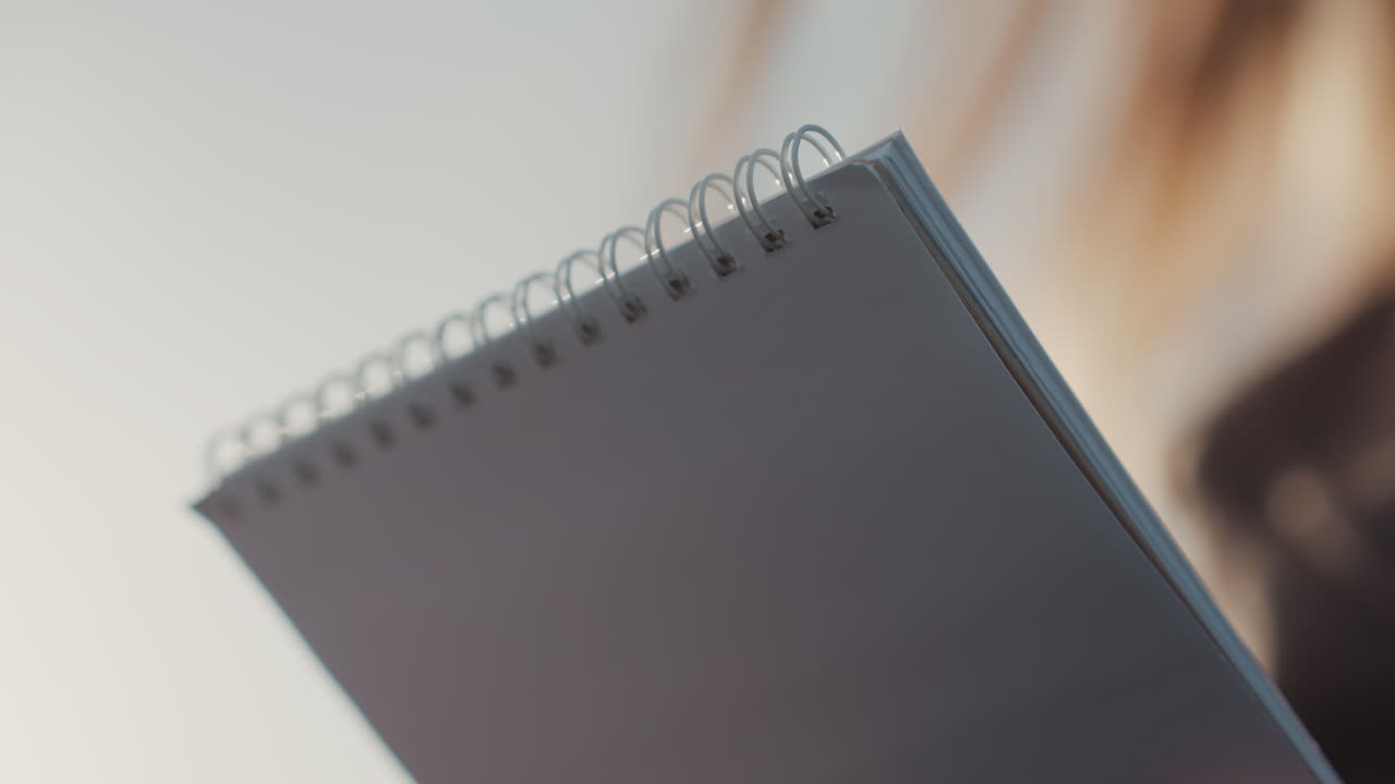 Woman examining spiral notebook closeup, designer reviewing layout ideas under soft natural light, hand flipping crisp pages, neutral palette, concentrated gesture, quiet workspace atmosphere