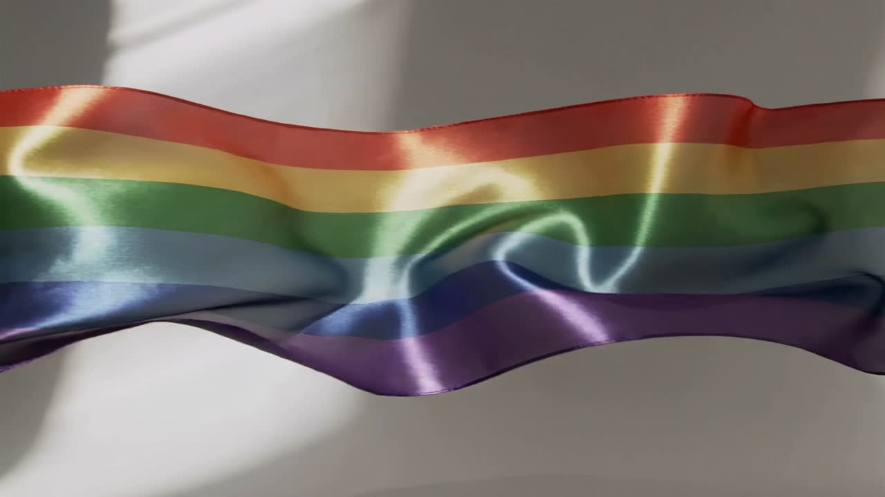 Bandiera arcobaleno di raso increspata che fluttua nell'aria fuori dall'inquadratura lungo la parete dello studio, catturando la luce e mostrando l'orlo