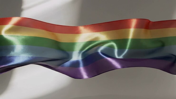 Bandiera arcobaleno di raso increspata che fluttua nell'aria fuori dall'inquadratura lungo la parete dello studio, catturando la luce e mostrando l'orlo