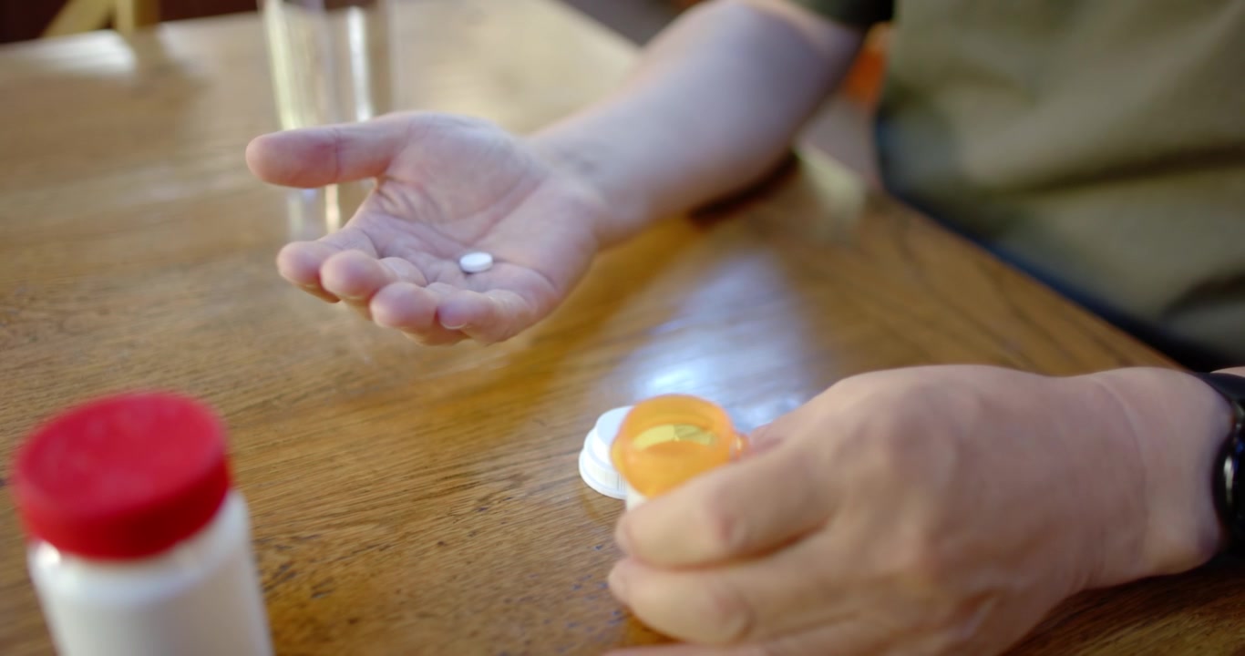Senior man at-home opening orange bottle cap, pouring tablets into left hand for meds near water