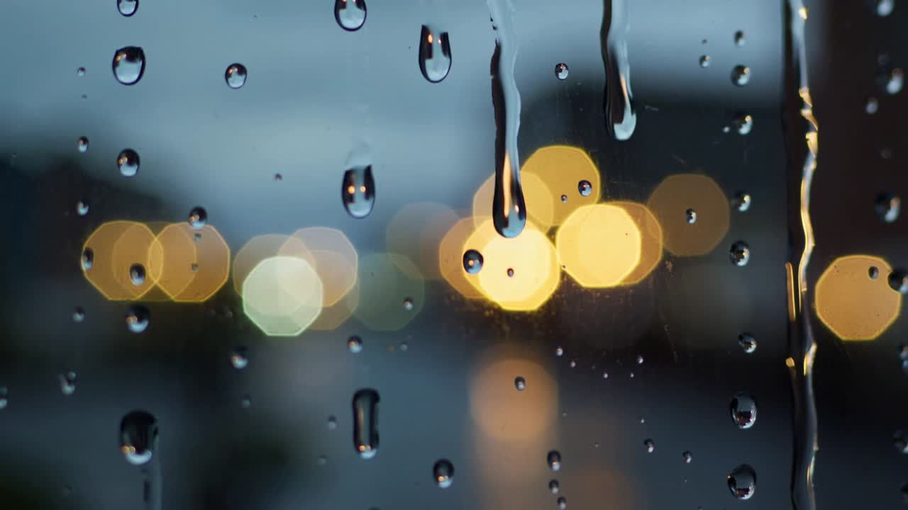 Showing gravity pulling raindrops into forming water streaks on window pane at dusk, bokeh lights