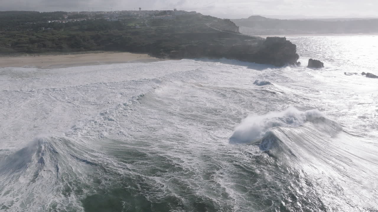 Wide aerial drone shot of big waves coming into shore on a day with giant waves in Nazaré, Portugal, Europe. Flying towards beach and people watching. Big wave surfing town