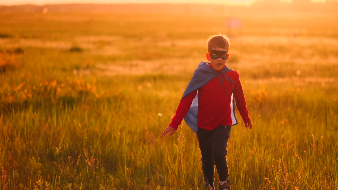 un niño con un traje y una máscara de superhéroe corre a través del campo al atardecer en la hierba soñando e imaginándose a sí mismo un héroe y un ganador. familia divertida y feliz