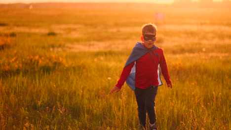 un niño con un traje y una máscara de superhéroe corre a través del campo al atardecer en la hierba soñando e imaginándose a sí mismo un héroe y un ganador. familia divertida y feliz