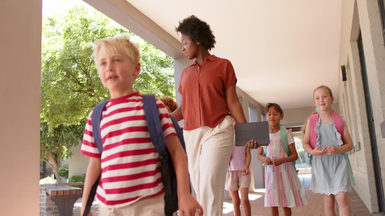 African American woman teacher leading kids along walkway with tablet urging blond boy to class