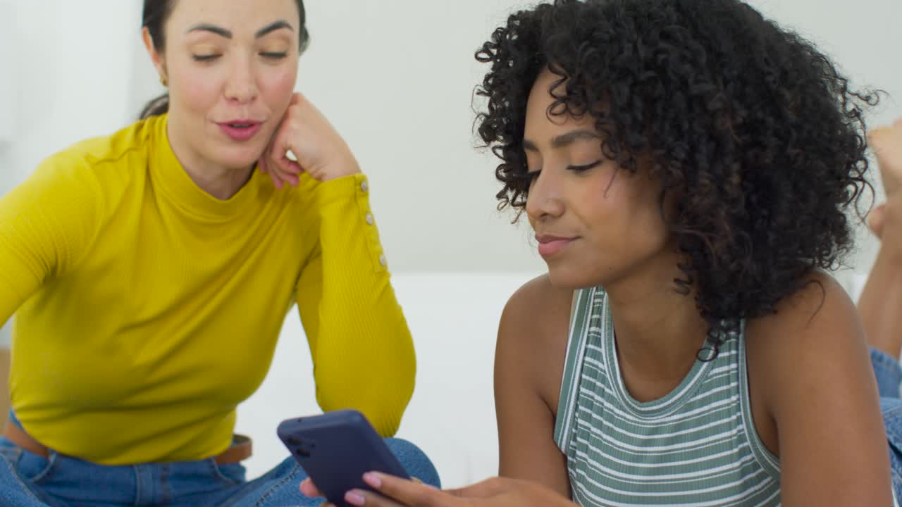 Diverse female friends leaning, yellow-top smiling while curly woman showing phone content on bed