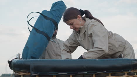 woman and man preparing hot air balloon basket outdoors adjusting ropes securing fuel tank and instrument bag wearing gloves under overcast sky