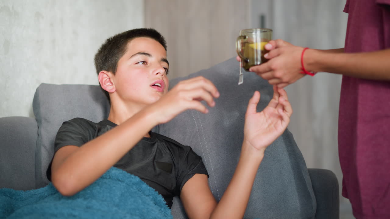 Sick child resting on couch collects tea from friend and drinks it for comfort. Friend has red lace bracelet on hand, showing care and affection in a warm home setting