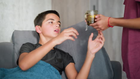 sick child resting on couch collects tea from friend and drinks it for comfort. friend has red lace bracelet on hand, showing care and affection in a warm home setting